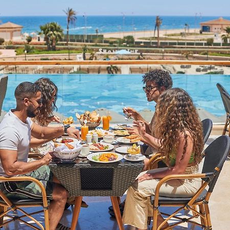 Tolip Resort El Galala Majestic Айн-Сохна Экстерьер фото The photo shows a group of five people enjoying a meal together outdoors, likely at a restaurant or cafe. They are seated at a table with a beautiful view of a swimming pool and the sea in the background. The table is filled with various dishes, and 