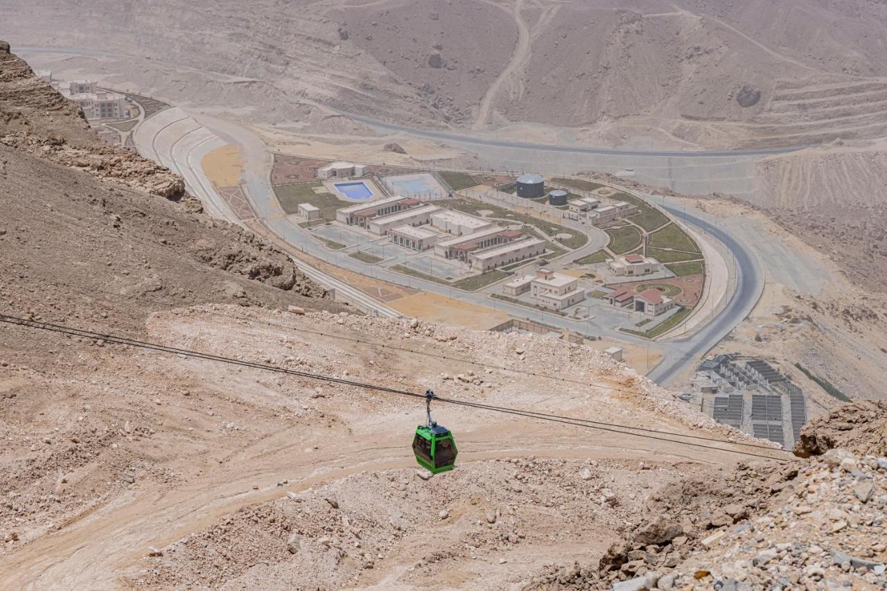 Tolip Resort El Galala Majestic Айн-Сохна Экстерьер фото The photo shows a mountainous landscape with a steep, dry terrain. In the foreground, there is a green cable car or gondola traveling along a cable. Below, a complex of buildings and roads can be seen, likely part of a resort or recreational area. Th