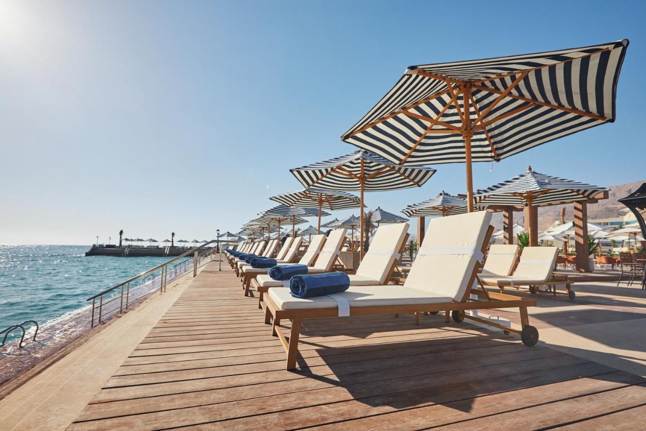 Tolip Resort El Galala Majestic Айн-Сохна Экстерьер фото The photo depicts a serene beachside setting featuring a row of sun loungers arranged on a wooden deck. Each lounger is equipped with a folded towel and shaded by striped umbrellas. In the background, the water sparkles under bright sunlight, and the