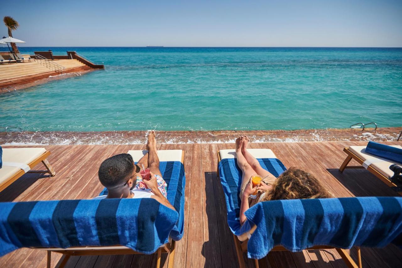 Tolip Resort El Galala Majestic Айн-Сохна Экстерьер фото The photo depicts two people relaxing on lounge chairs by the waterfront. They are positioned on a wooden deck with a view of the clear, turquoise water. Both individuals are reclining, enjoying the sun, and each has a drink in hand. The scene convey