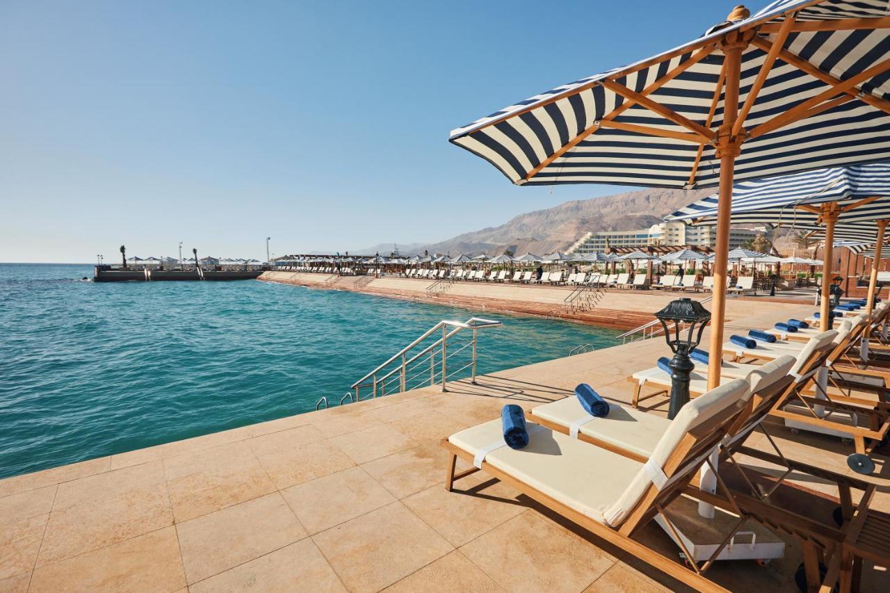 Tolip Resort El Galala Majestic Айн-Сохна Экстерьер фото The photo shows a scenic beachside area with clear blue water. In the foreground, there are several sun loungers set up on a tiled surface, with blue towels neatly rolled on each one. The loungers are shaded by striped umbrellas. In the distance, a w